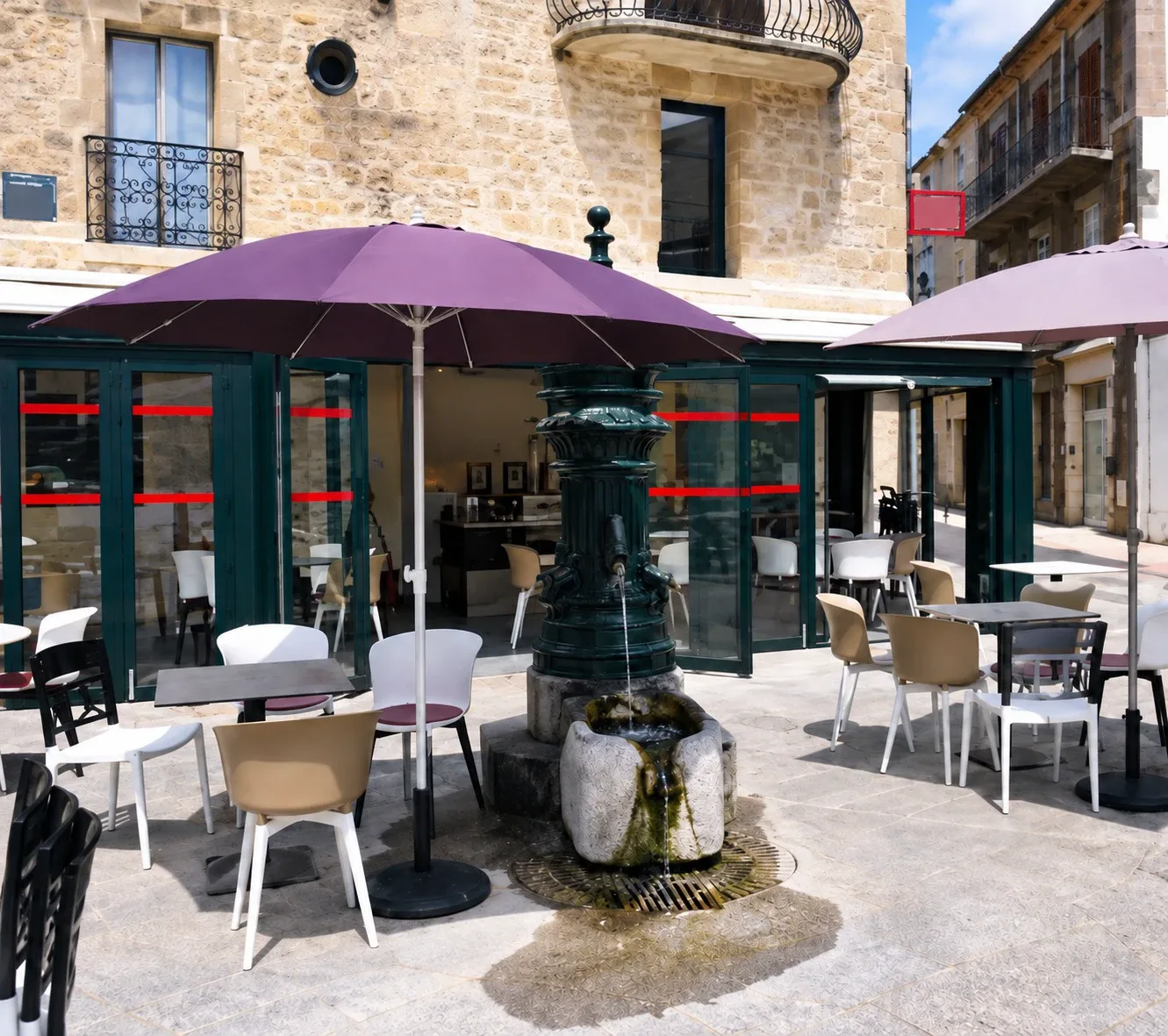 Le bar-restaurant sur sa place&nbsp;: terrasse sous parasols autour de la fontaine, façade vitrée au pied de l’immeuble en pierre