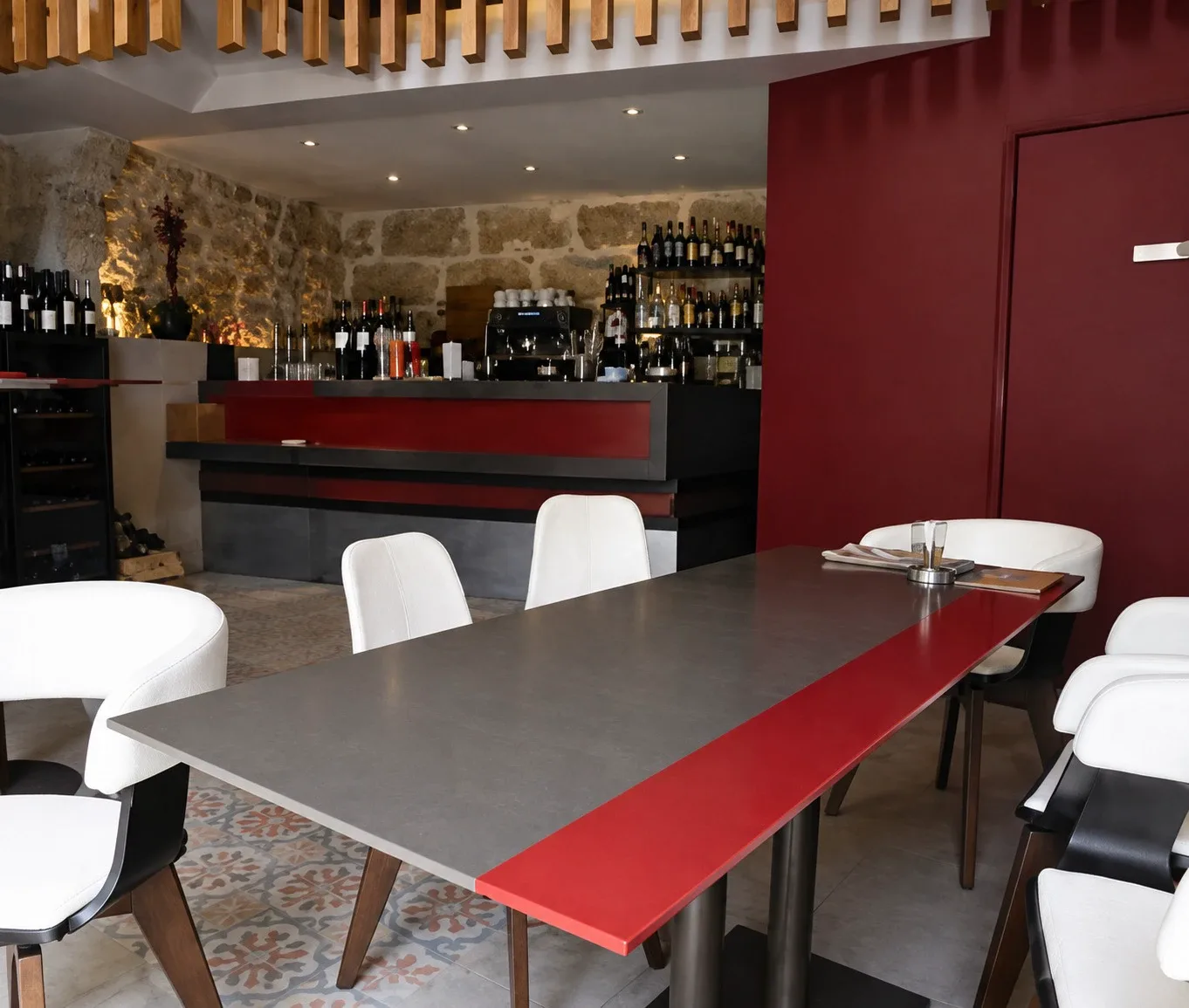 La salle livrée&nbsp;: grande table grise à bande rouge, chaises blanches, comptoir et mur de pierre éclairé au fond