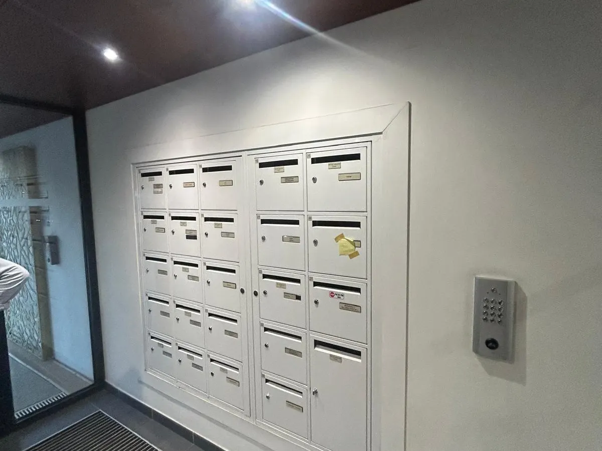 Photographie du mur des boîtes aux lettres avant travaux : boîtes blanches sur mur blanc, interphone, éclairage ponctuel