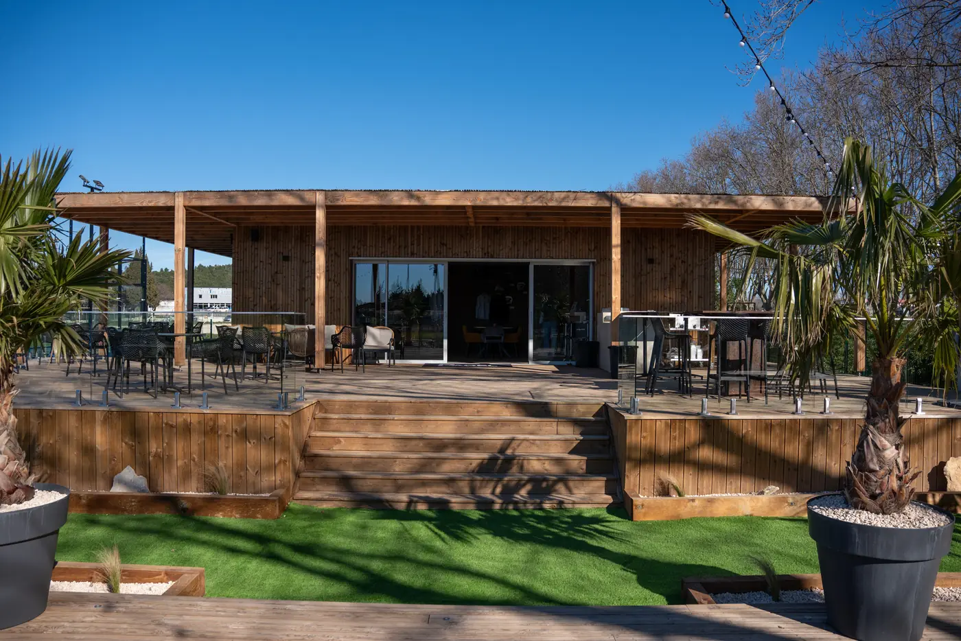 Le club-house en bois, sa terrasse sur pilotis et son escalier, entre deux palmiers