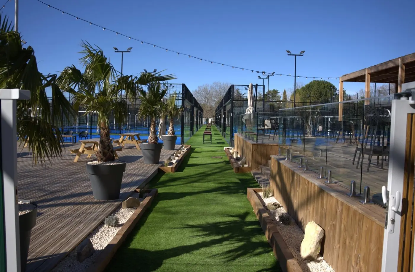Allée en gazon bordée de palmiers en pot, entre les terrasses en bois et les terrains de padel vitrés